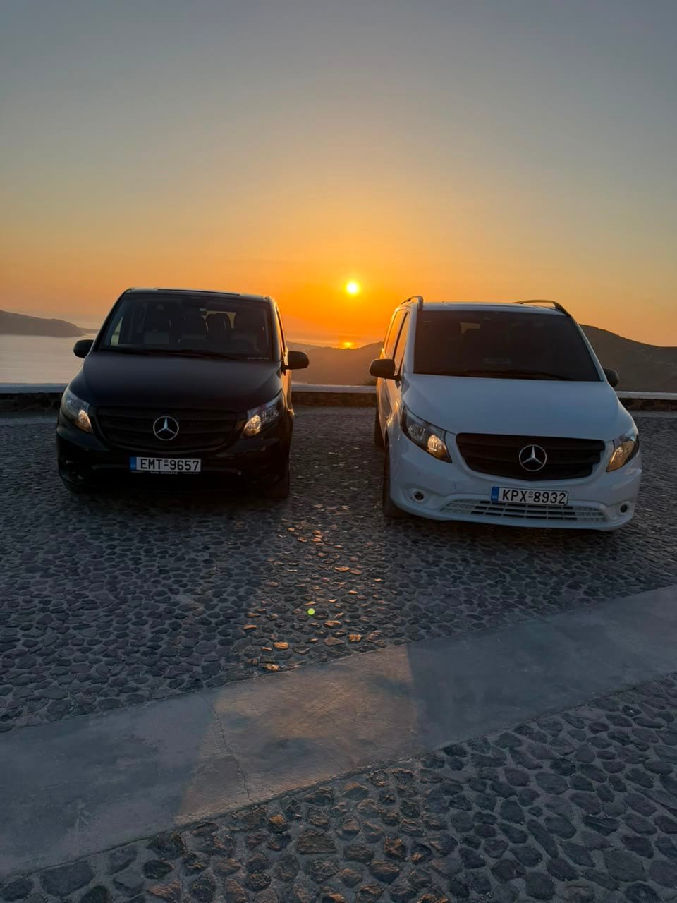 The two Ride with Dimitris vans — a black and a white Mercedes-Benz Vito — parked at a Santorini sunset viewpoint over the caldera